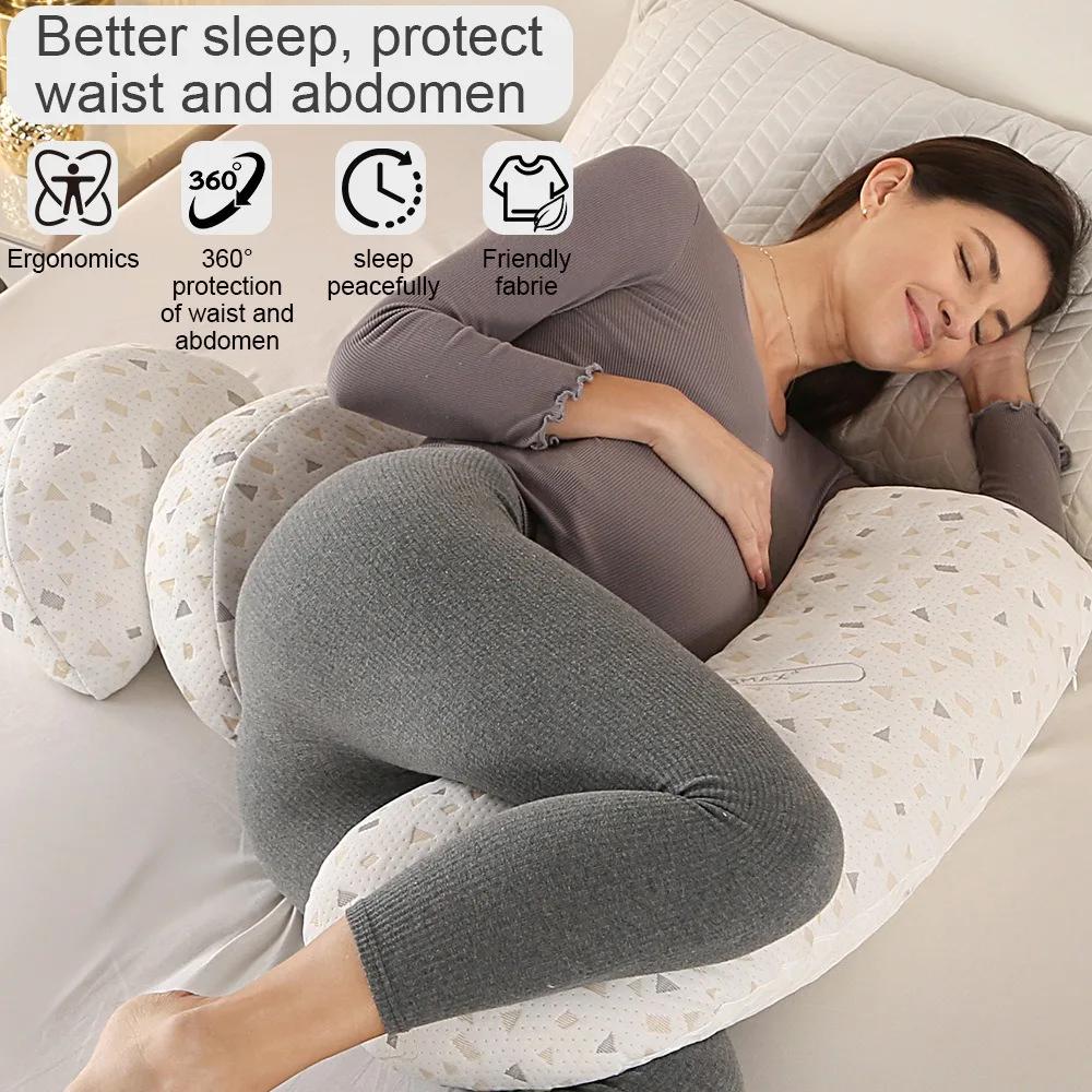 A pregnant woman reclining on a white U-shaped maternity pillow with geometric patterns, wearing a gray long-sleeve top and gray leggings. The image includes text overlays highlighting benefits such as ergonomic support, 360-degree protection, peaceful sleep, and friendly fabric.